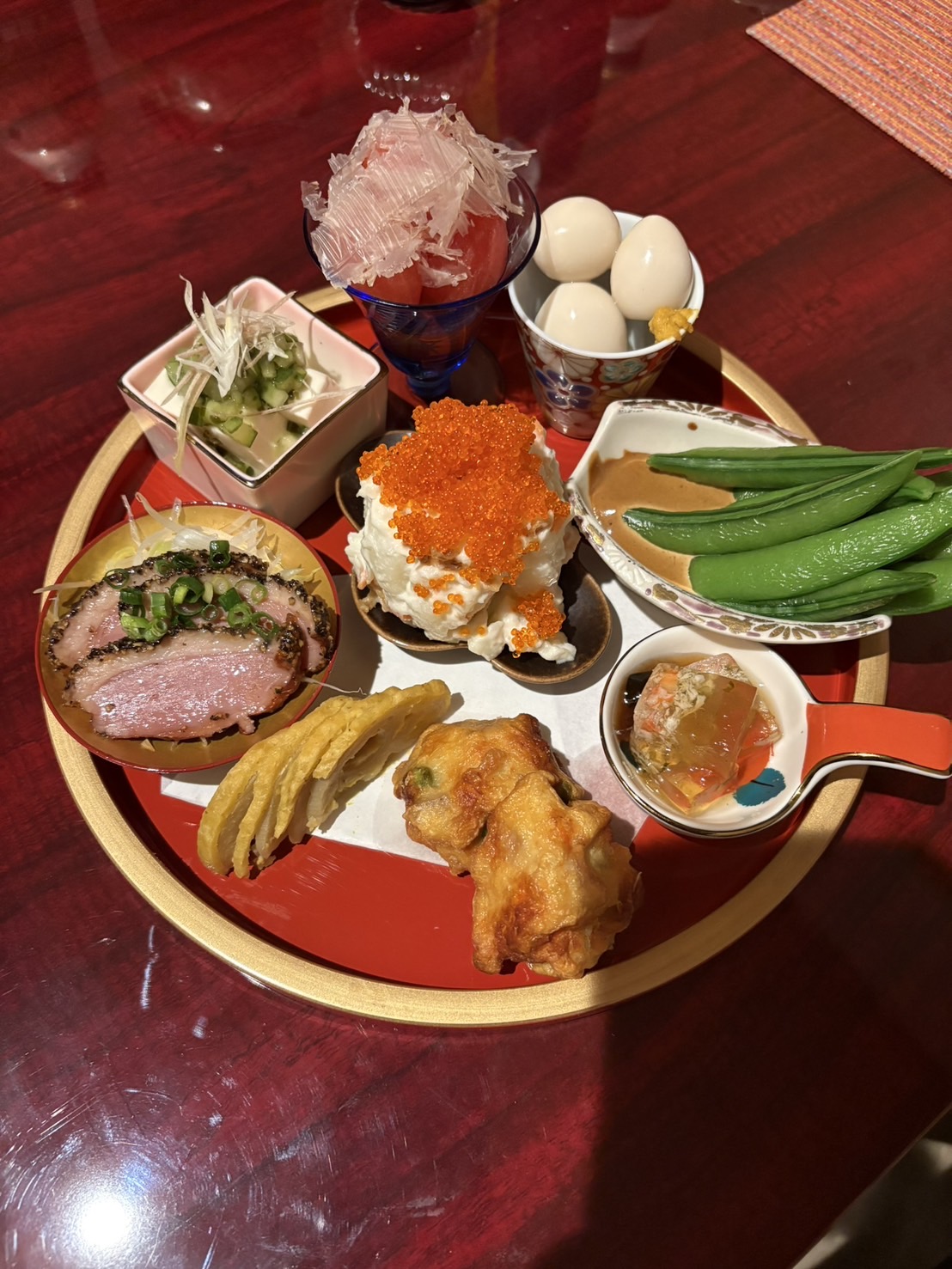 赤い円形トレイに和食の前菜、卵、いくら、豆腐、野菜、揚げ物、飲み物が並ぶ。